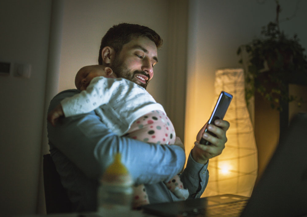 Father holding sleeping baby while using phone at night